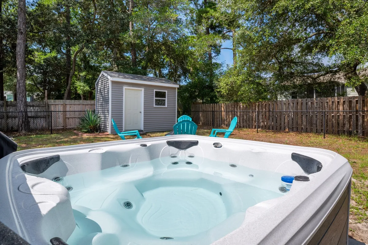 This shot captures a peaceful part of the backyard with a hot tub, vibrant blue chairs, a visible storage shed, and mature trees providing shade. The scene feels tranquil and social, perfect for relaxing, stargazing, or gathering after a day at the beach.