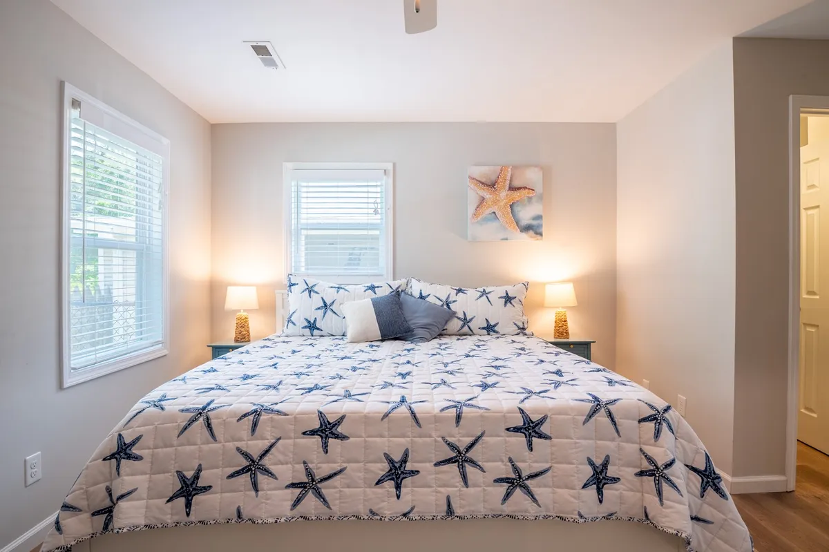 This image showcases a serene bedroom with a bed dressed in a starfish-patterned quilt, decorative lamps, starfish wall art, and plenty of natural light streaming in through the windows. The room is bright, tranquil, and evokes a coastal atmosphere perfect for relaxation.