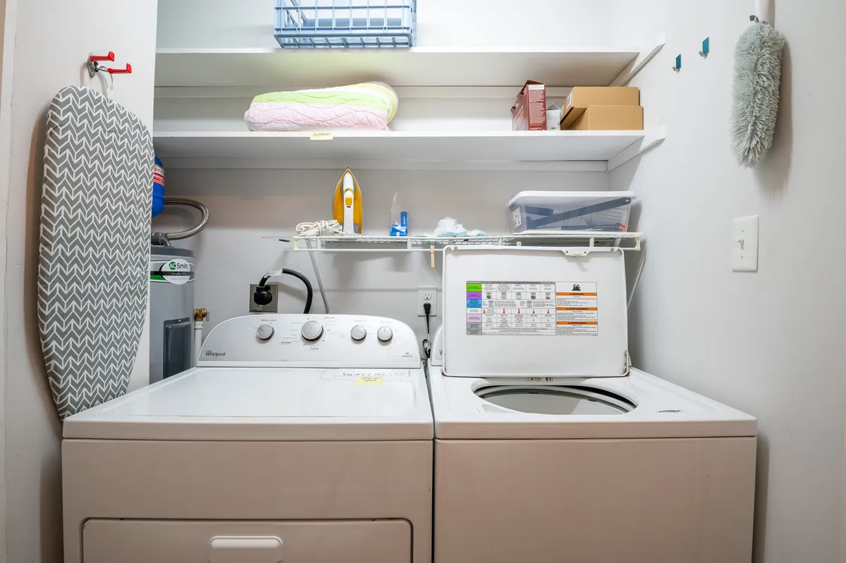 A laundry area featuring a washing machine and dryer side-by-side, with an ironing board and shelves for storage. The scene is practical, well-organized, and brightly lit, emphasizing ease of daily routines and in-home accessibility.