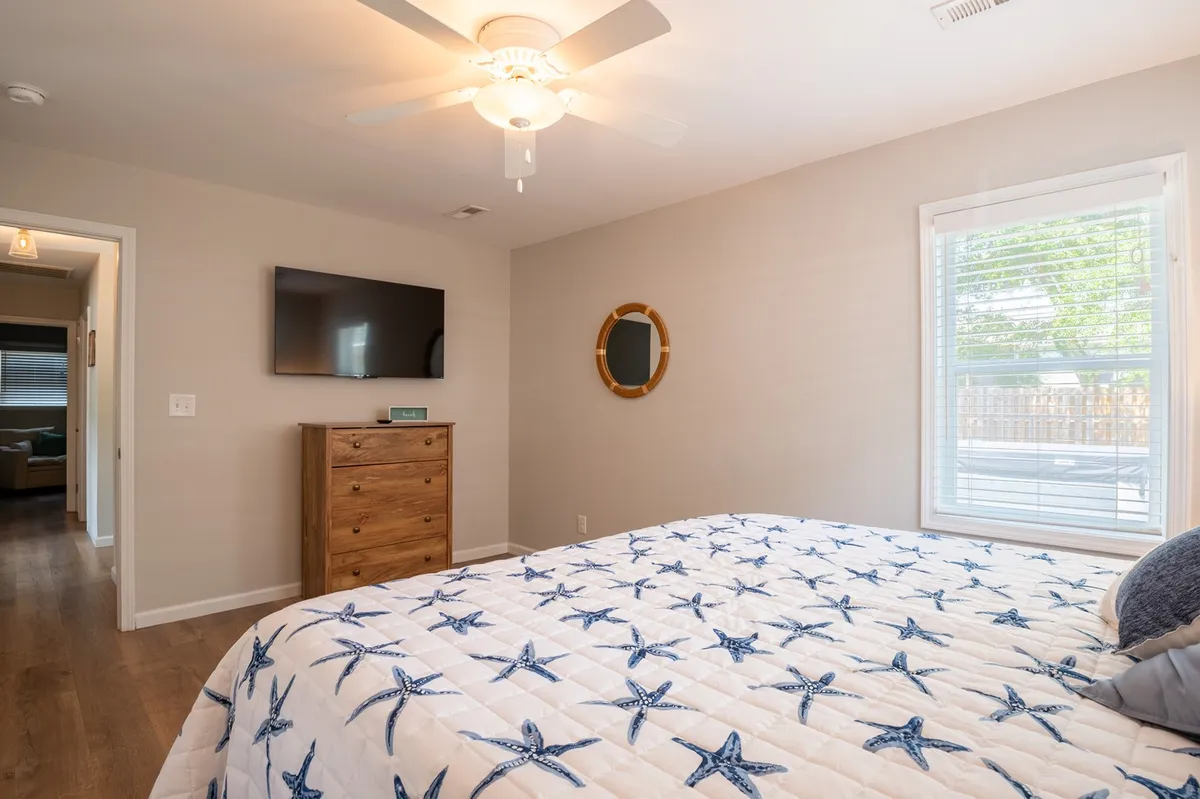 A cozy bedroom with starfish-patterned bedding, wooden dresser, wall-mounted TV, round mirror, and a window letting in natural light. The room is uncluttered and accessible, designed for restful, easy navigation.