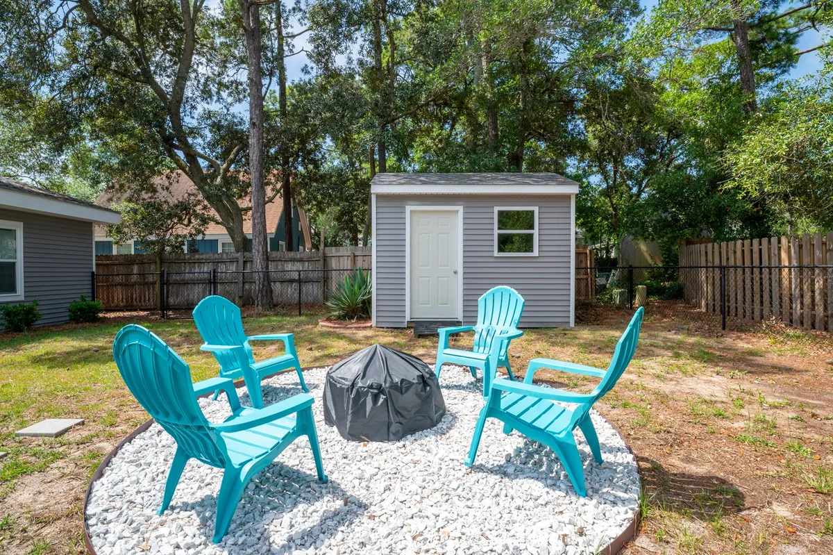An inviting backyard scene featuring bright turquoise chairs arranged around a covered fire pit, with a small storage shed in the background. The setting suggests a welcoming space for outdoor relaxation, evening gatherings, and family activities under open skies.