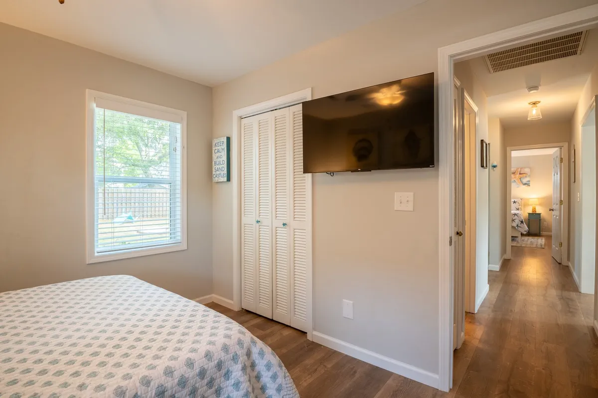 A comfortable bedroom scene featuring a bed with patterned bedding, a wall-mounted television, a window letting in natural light, and closet doors. The room appears spacious and offers a cozy space for unwinding or watching TV in bed.